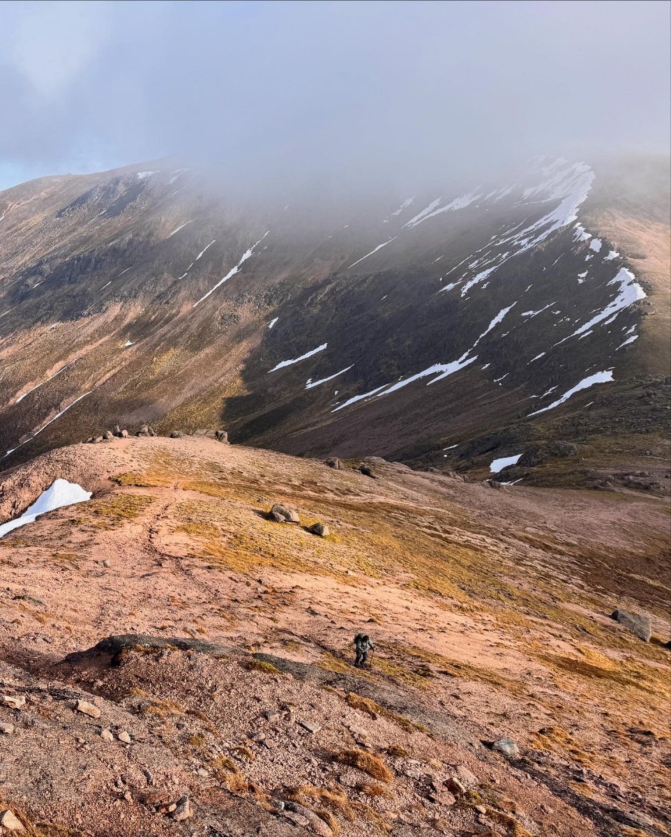 Solo hiker ascending a mountain ridge with misty peaks and patches of snow in the distance